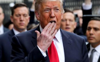 Donald Trump in a dark suit and red tie with his hand near his mouth, surrounded by men