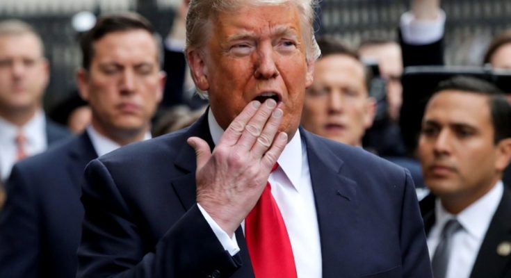 Donald Trump in a dark suit and red tie with his hand near his mouth, surrounded by men