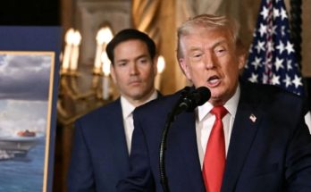 Donald Trump speaking at a podium with a microphone, wearing a dark suit and red tie