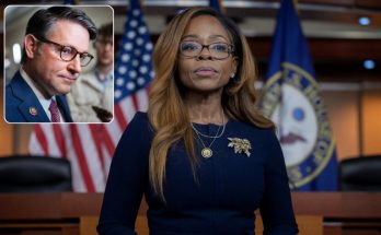 A woman with long brown hair wearing glasses and a dark blue dress stands in front of