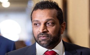 Kash Patel in dark suit with red tie, looking to his right in indoor hallway setting