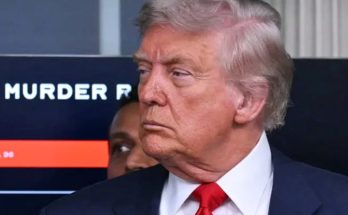 Donald Trump in a dark suit and red tie, with a screen behind him displaying partial