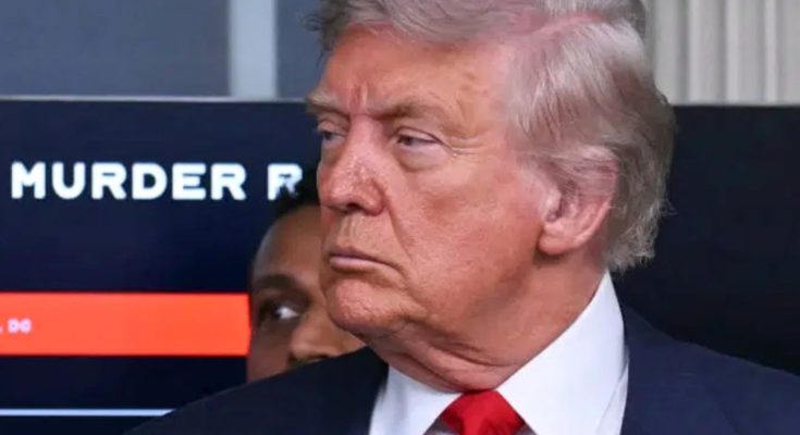 Donald Trump in a dark suit and red tie, with a screen behind him displaying partial