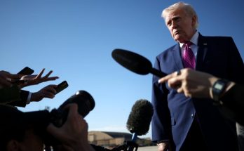 Donald Trump in a dark blue suit and magenta tie stands outdoors under clear blue sky