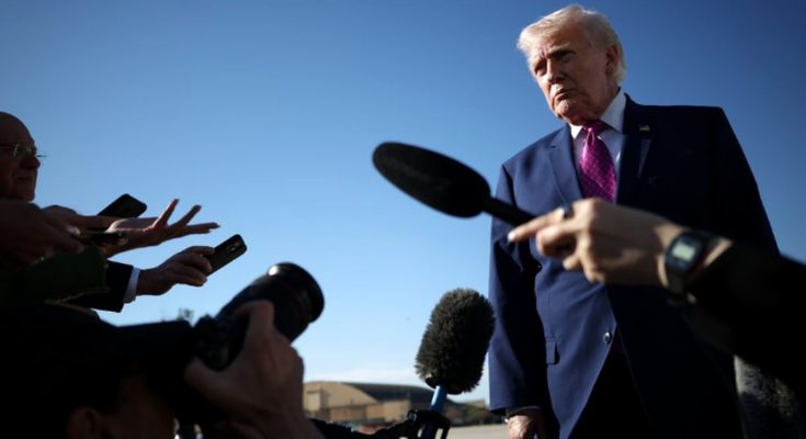 Donald Trump in a dark blue suit and magenta tie stands outdoors under clear blue sky