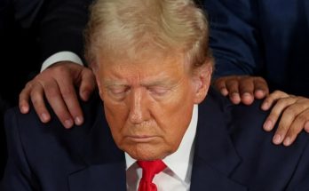 Donald Trump with eyes closed and head bowed while two pairs of hands rest on his