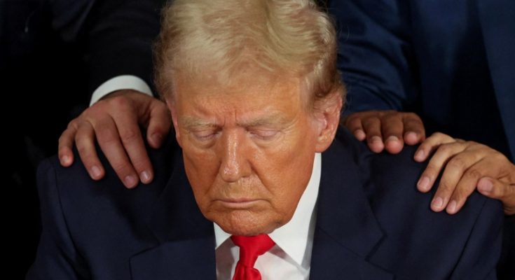 Donald Trump with eyes closed and head bowed while two pairs of hands rest on his