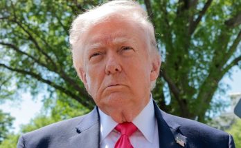 Donald Trump wearing a dark suit, white shirt, and red tie with an American flag pin