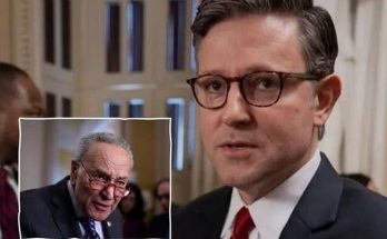 A man in a dark suit with a red tie and glasses stands in a hallway, with an inset image