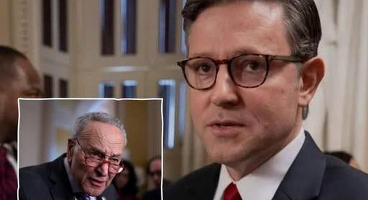 A man in a dark suit with a red tie and glasses stands in a hallway, with an inset image