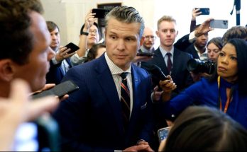 Pete Hegseth in a navy pinstripe suit surrounded by reporters and microphones in a