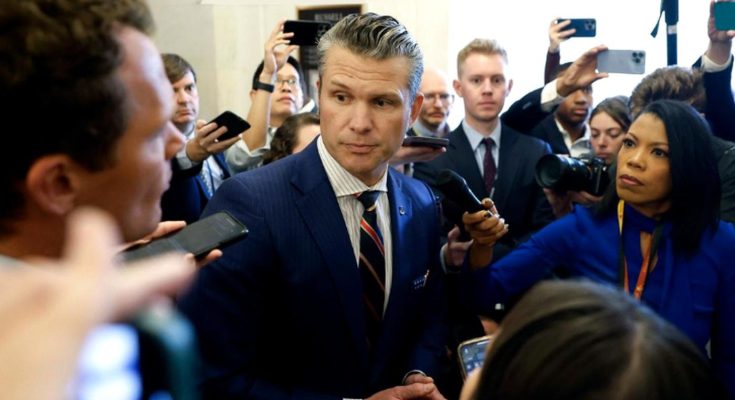 Pete Hegseth in a navy pinstripe suit surrounded by reporters and microphones in a