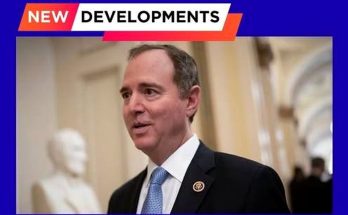 A man in a dark suit with a light blue tie and congressional pin stands in a hallway