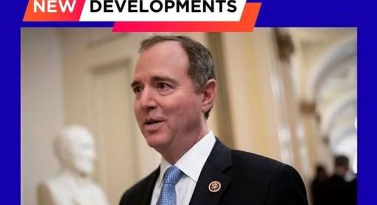 A man in a dark suit with a light blue tie and congressional pin stands in a hallway