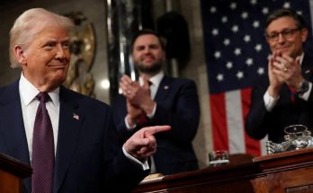 Donald Trump in a dark suit and red tie pointing with his right hand while two men in