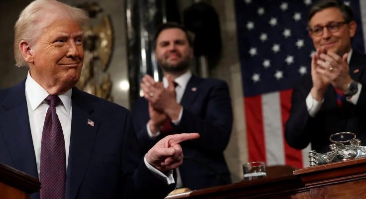 Donald Trump in a dark suit and red tie pointing with his right hand while two men in