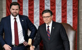 Two men in suits stand in front of an American flag backdrop. The man on the left wears
