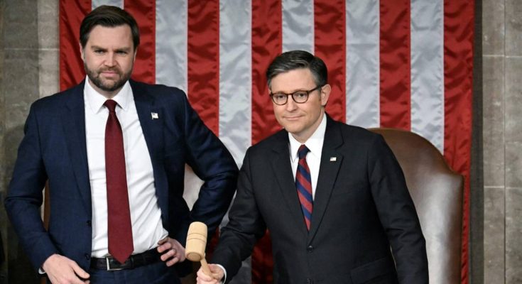 Two men in suits stand in front of an American flag backdrop. The man on the left wears