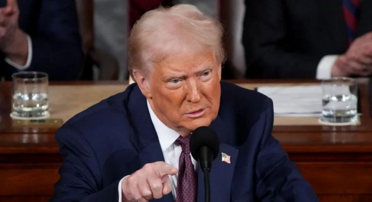 Donald Trump speaking at a podium with a microphone, wearing a dark blue suit and red