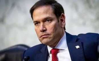 Marco Rubio in a navy suit and red tie seated at a hearing with a microphone