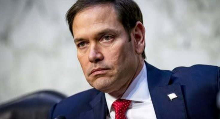 Marco Rubio in a navy suit and red tie seated at a hearing with a microphone