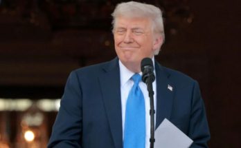 A man in a dark suit and blue tie standing at a podium with a microphone, holding
