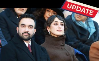 A man in a black coat and red tie sits next to a woman in a brown high-collared coat