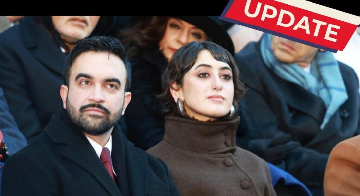 A man in a black coat and red tie sits next to a woman in a brown high-collared coat