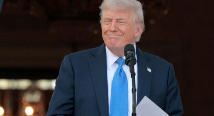 A man in a dark suit and blue tie standing at a podium with a microphone, holding
