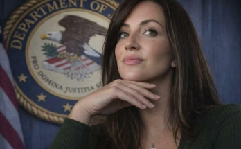 A woman with long brown hair wearing a green sweater and cross necklace rests her chin