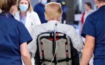 A person with blonde hair wearing a white shirt sits in a wheelchair with a medical tube
