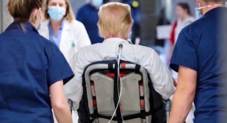 A person with blonde hair wearing a white shirt sits in a wheelchair with a medical tube