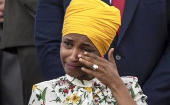 ilhan Omar wearing yellow turban and floral blouse wipes away tears with ringed hand