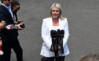 woman in white blazer speaks to media with microphones outdoors