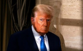 donald Trump stands in a navy suit and bright blue tie against a travertine column and