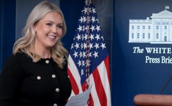 woman smiling at White House press briefing podium with flag backdrop