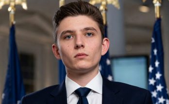 young man in suit with flags in background, serious expression, political setting