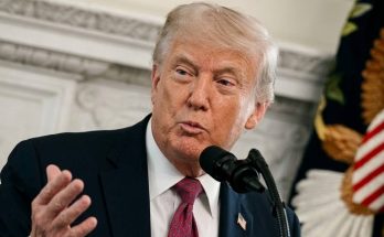 donald Trump speaking at podium with microphone, wearing dark suit and red patterned tie