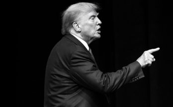 man in suit gestures emphatically on stage, black background