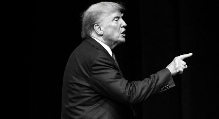 man in suit gestures emphatically on stage, black background