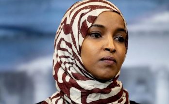 woman in patterned hijab stands against blurred background with American flag