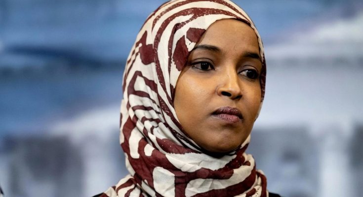 woman in patterned hijab stands against blurred background with American flag