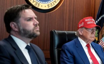two men at a conference table, one wearing a red cap, serious expressions