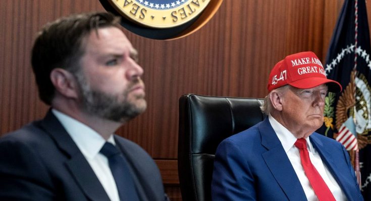 two men at a conference table, one wearing a red cap, serious expressions
