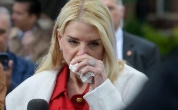 woman in red coat wiping nose amid crowd during press event