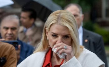 blonde woman in red coat speaks at outdoor press event