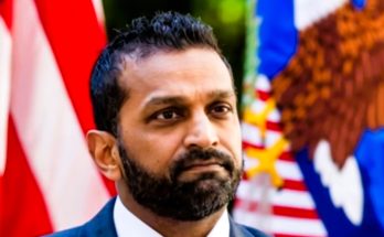 serious man stands before American flags outdoors