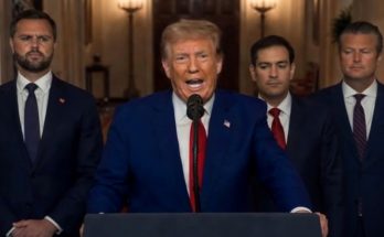 politician delivers speech flanked by serious men in suits